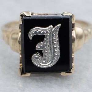 May include: A gold-toned ring featuring a rectangular black onyx stone. The stone is adorned with a silver-colored letter "J" in an ornate, stylized font. The ring's band has a textured design.