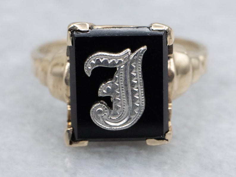 May include: A gold-toned ring featuring a rectangular black onyx stone. The stone is adorned with a silver-colored letter "J" in an ornate, stylized font. The ring's band has a textured design.