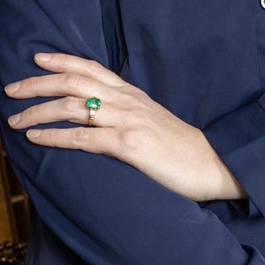 May include: A gold ring with a large, square-cut emerald gemstone and smaller accent stones. The ring is set against a dark blue background, with the hand and arm of a person wearing a dark blue shirt.