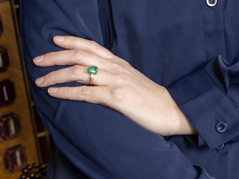 May include: A gold ring with a large, square-cut emerald gemstone and smaller accent stones. The ring is set against a dark blue background, with the hand and arm of a person wearing a dark blue shirt.