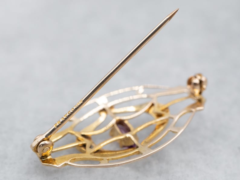 May include: A gold-toned brooch with a delicate, openwork design featuring a purple gemstone. The brooch has a pin and catch closure.