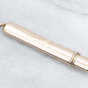 May include: A gold-colored, cylindrical container with a loop at one end. The container has a decorative, engraved design on its surface. The object is likely a vintage or antique item.