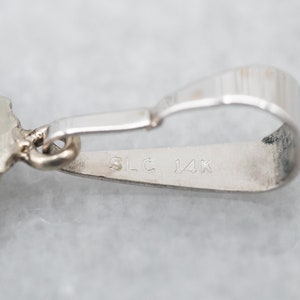 May include: Close-up of a silver pendant with a detailed design. The pendant has a loop for attaching a chain and is marked "SLC 14K" on the back.
