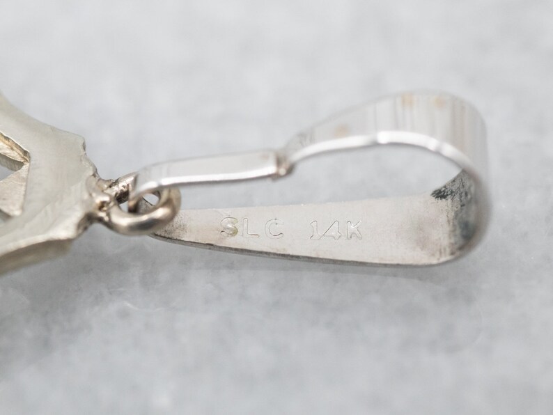 May include: Close-up of a silver pendant with a detailed design. The pendant has a loop for attaching a chain and is marked "SLC 14K" on the back.