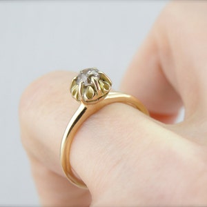 Puede incluir: Un anillo de oro con una sola piedra preciosa redonda y transparente engastada en garras. El anillo est&aacute; en un dedo.