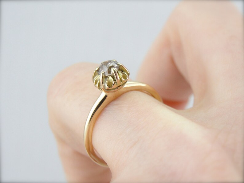 Puede incluir: Un anillo de oro con una sola piedra preciosa redonda y transparente engastada en garras. El anillo est&aacute; en un dedo.