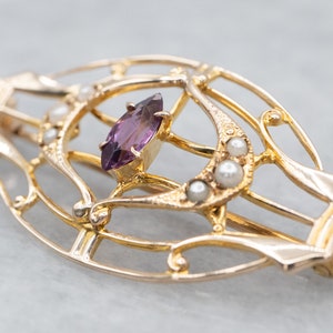 May include: A gold-toned brooch with a delicate, intricate design featuring a purple gemstone and pearl accents.