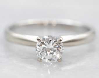 Puede incluir: Un anillo de compromiso solitario simple con un diamante redondo brillante engastado en una banda de oro blanco.