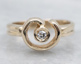 May include: A gold ring with a unique design. The ring features a central, clear gemstone set within a swirling, openwork design. The band is a simple, polished gold. The ring is set against a soft, neutral background.