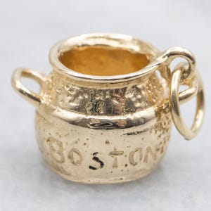 May include: A gold-colored charm in the shape of a small pot with two handles. The word "BOSTON" is engraved on the side. The charm has a textured surface and a loop for attaching to a chain or bracelet.