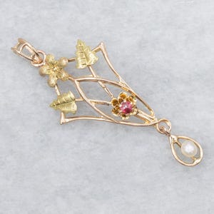Puede incluir: Un colgante dorado con detalles florales y de hojas, con una gema rosa central y una perla colgante. El colgante tiene un diseño delicado y ornamentado.