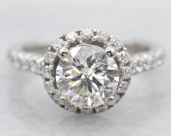May include: A silver engagement ring with a large, round, clear diamond center stone surrounded by a halo of smaller diamonds. The band is also set with smaller diamonds.