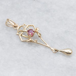Puede incluir: Colgante dorado con una gema rosa redonda en el centro. El colgante presenta un diseño calado con elementos geométricos y florales, incluyendo un colgante en forma de lágrima en la parte inferior. El colgante es probablemente para un collar.