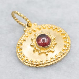 May include: Gold pendant with a teardrop shape, featuring a central, round, dark red gemstone. The pendant has a textured surface with a beaded border and a twisted bail. A sunburst design surrounds the gemstone.