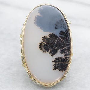 May include: A gold ring featuring a large oval dendritic agate stone. The stone is white with black dendritic patterns.