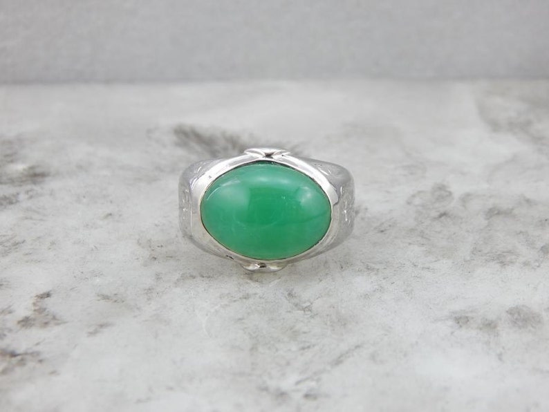 May include: A silver ring with a green oval gemstone set in the center. The ring has a textured band.