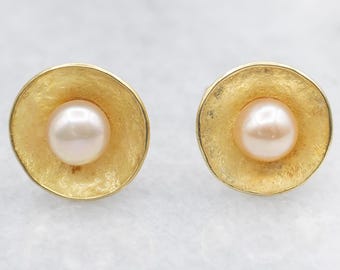 May include: A pair of gold-toned stud earrings. Each earring features a light pink pearl set in a textured, circular gold setting. The earrings are displayed against a light background, highlighting their design.