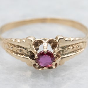 Puede incluir: Un anillo de oro con una gema redonda de color rosa intenso en el centro. El anillo tiene un diseño ornamentado con elementos decorativos en la banda y alrededor del engaste de la gema. El anillo está sobre un fondo claro.