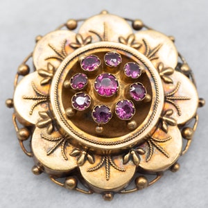 May include: A gold-toned brooch with a floral design and a central circle set with pink stones.