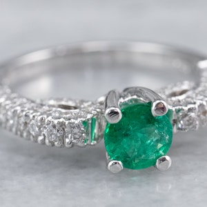 May include: A silver ring featuring a large emerald green gemstone surrounded by smaller diamonds. The ring is set in a delicate, intricate design.