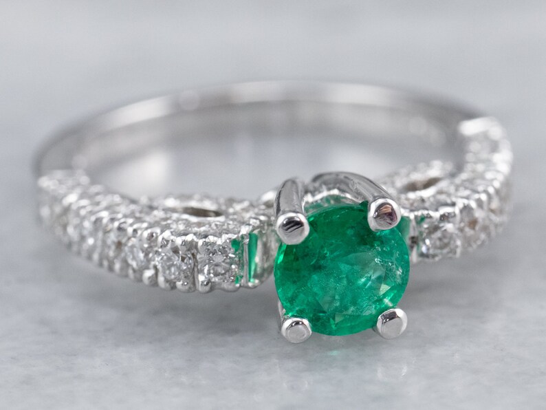May include: A silver ring featuring a large emerald green gemstone surrounded by smaller diamonds. The ring is set in a delicate, intricate design.