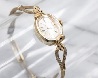 May include: A vintage gold-toned wristwatch with an oval face and a mesh band. The watch features a delicate design with a bow detail at the top and bottom of the watch face. The watch hands point to 10:09.