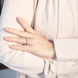 K&ouml;nnte beinhalten: Ein silberner Verlobungsring mit einem gro&szlig;en, runden, klaren Edelstein wird auf einer Hand gezeigt. Der Ring ist auf einem zarten Band mit kleineren Akzentsteinen gefasst. Die Person tr&auml;gt eine hellrosa Langarmbluse.