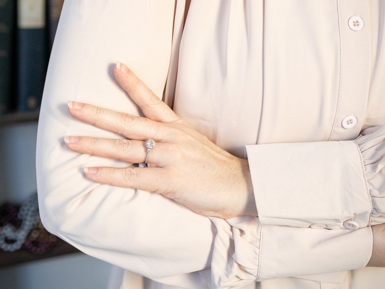 K&ouml;nnte beinhalten: Ein silberner Verlobungsring mit einem gro&szlig;en, runden, klaren Edelstein wird auf einer Hand gezeigt. Der Ring ist auf einem zarten Band mit kleineren Akzentsteinen gefasst. Die Person tr&auml;gt eine hellrosa Langarmbluse.