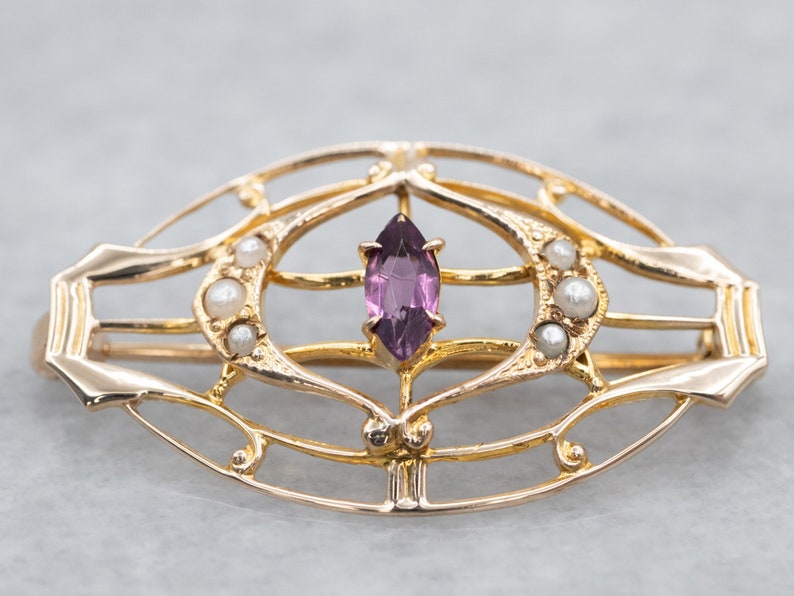 May include: A gold-toned brooch with a delicate, openwork design featuring a purple gemstone and small pearls.