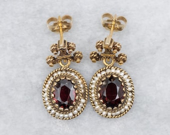 May include: A pair of gold-toned drop earrings. Each earring features an oval, deep red gemstone surrounded by small white pearls and a twisted gold rope design. The earrings have a decorative top with a post back.