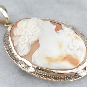 May include: A cameo pendant featuring a carved shell with a woman's profile and floral details. The pendant is set in a gold filigree frame.