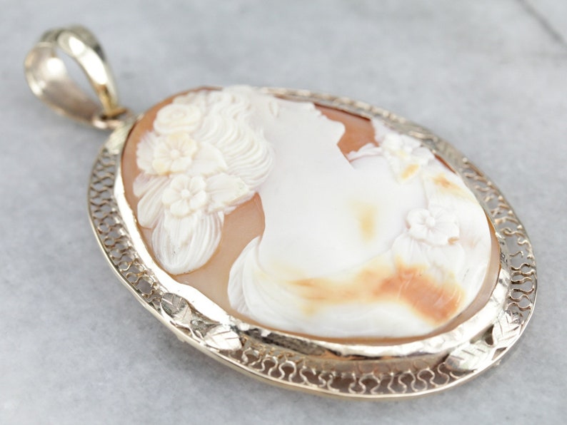 May include: A cameo pendant featuring a carved shell with a woman's profile and floral details. The pendant is set in a gold filigree frame.