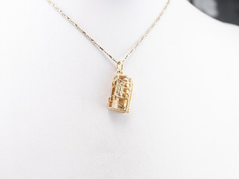 May include: A gold chain necklace with a pendant shaped like a small, rectangular box with a detailed design. The box has a small, clear stone in the center.
