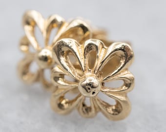 以下が含まれることがあります： 2つの金色の花の形をしたイヤリングのクローズアップ。各イヤリングは、6つの湾曲した花びらに囲まれた中央の球体を特徴としています。イヤリングは、磨かれた反射面を持っており、貴金属で作られていることを示唆しています。