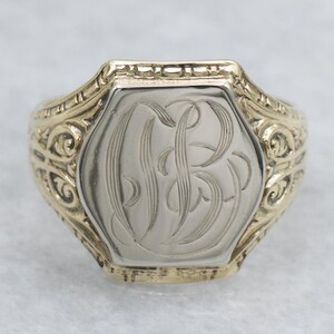May include: A gold and silver signet ring with an engraved monogram. The ring features a rectangular face with a detailed, ornate gold band. The silver face displays an engraved monogram, showcasing a classic and elegant design.