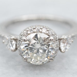 K&ouml;nnte beinhalten: Ein Diamant-Verlobungsring mit einem gro&szlig;en, rund geschliffenen Mittelstein, der von kleineren Diamanten umgeben ist. Der Ring hat ein Band aus Wei&szlig;gold oder Platin und zus&auml;tzliche Diamanten an den Seiten.