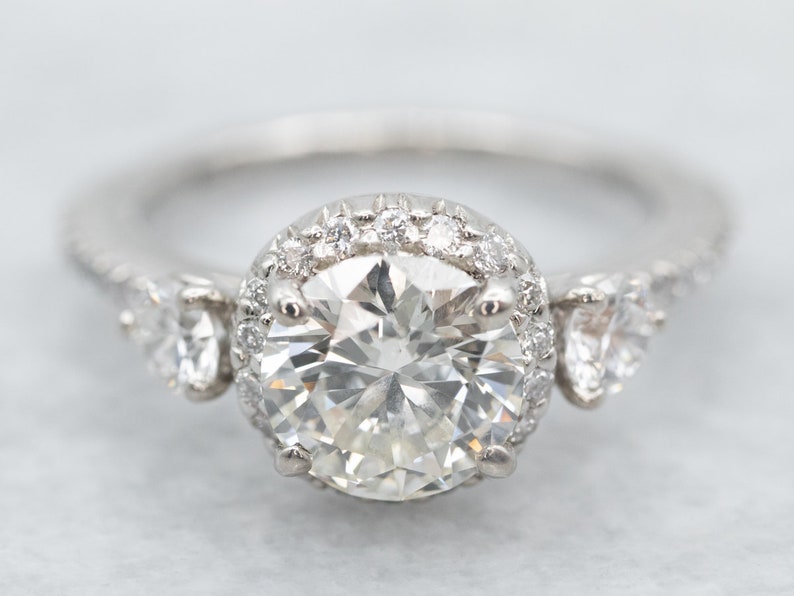 K&ouml;nnte beinhalten: Ein Diamant-Verlobungsring mit einem gro&szlig;en, rund geschliffenen Mittelstein, der von kleineren Diamanten umgeben ist. Der Ring hat ein Band aus Wei&szlig;gold oder Platin und zus&auml;tzliche Diamanten an den Seiten.