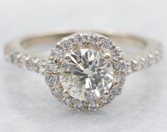 May include: A round-cut diamond engagement ring with a halo of smaller diamonds. The ring has a gold band with additional small diamonds set along the band. The ring is set against a white background.