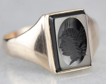 May include: A gold ring set with a black onyx stone, featuring a carved profile of a person with a feathered headdress.