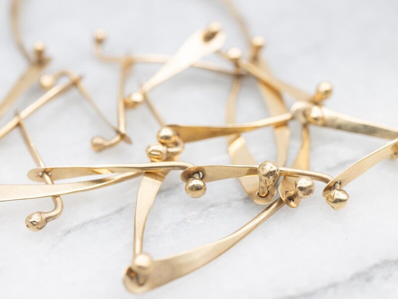 May include: Close-up of a gold-toned necklace with an abstract design. The necklace features multiple interconnected, curved elements with small, spherical accents. The metal has a polished finish, and the piece is set against a light background.
