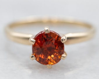 Puede incluir: Un anillo de oro con una piedra preciosa redonda de color naranja engastada en un engaste de cuatro garras.
