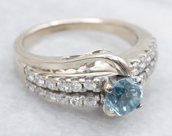 May include: A white gold ring set with a round, light blue gemstone and surrounded by small, clear gemstones. The ring has a unique, flowing design with multiple bands.