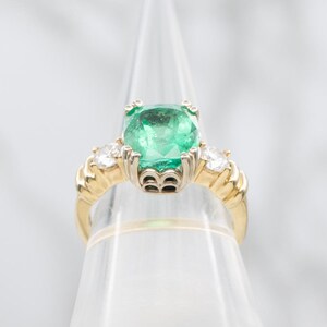 May include: A gold ring featuring a large, square-cut emerald center stone, flanked by two smaller diamonds. The ring has a detailed, textured band and is displayed on a clear stand. The emerald is a vibrant green color.