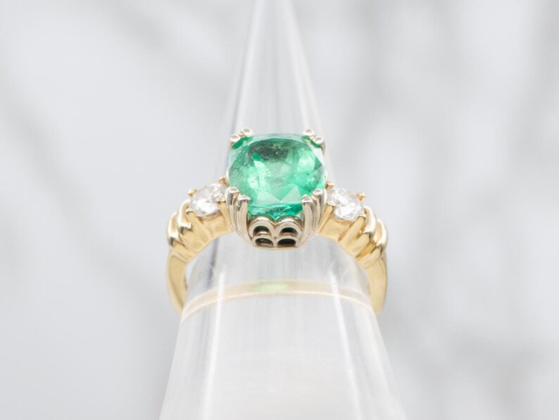 May include: A gold ring featuring a large, square-cut emerald center stone, flanked by two smaller diamonds. The ring has a detailed, textured band and is displayed on a clear stand. The emerald is a vibrant green color.