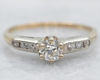 May include: A vintage engagement ring with a single round diamond set in a platinum band. The band is accented with a row of smaller diamonds on each side.