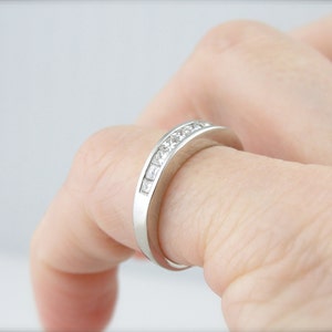 May include: A silver band with a row of small, square-shaped diamonds set in a channel setting.