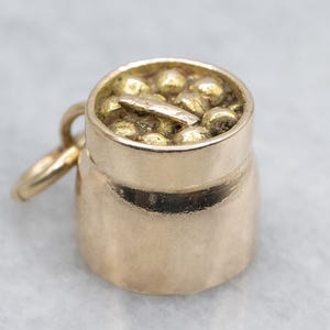 Könnte beinhalten: Ein goldfarbener Anhänger in Form eines kleinen Behälters, möglicherweise ein Topf oder eine Tasse, gefüllt mit kleinen, runden, goldfarbenen Objekten. Ein kleines, flaches, goldfarbenes Objekt befindet sich ebenfalls darin. Der Anhänger hat eine Schlaufe zum Befestigen an einer Kette.