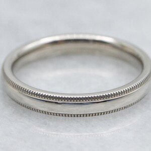 May include: A close-up of a silver ring with a detailed, textured design. The ring has a smooth, polished center band, bordered by a decorative, ridged pattern. The ring is likely made of a precious metal, such as silver or platinum.