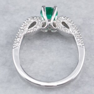 May include: A silver ring featuring a large emerald green gemstone surrounded by smaller diamonds. The ring has a delicate, intricate design.
