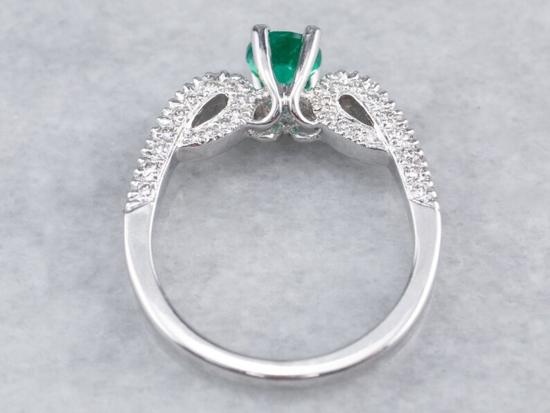 May include: A silver ring featuring a large emerald green gemstone surrounded by smaller diamonds. The ring has a delicate, intricate design.